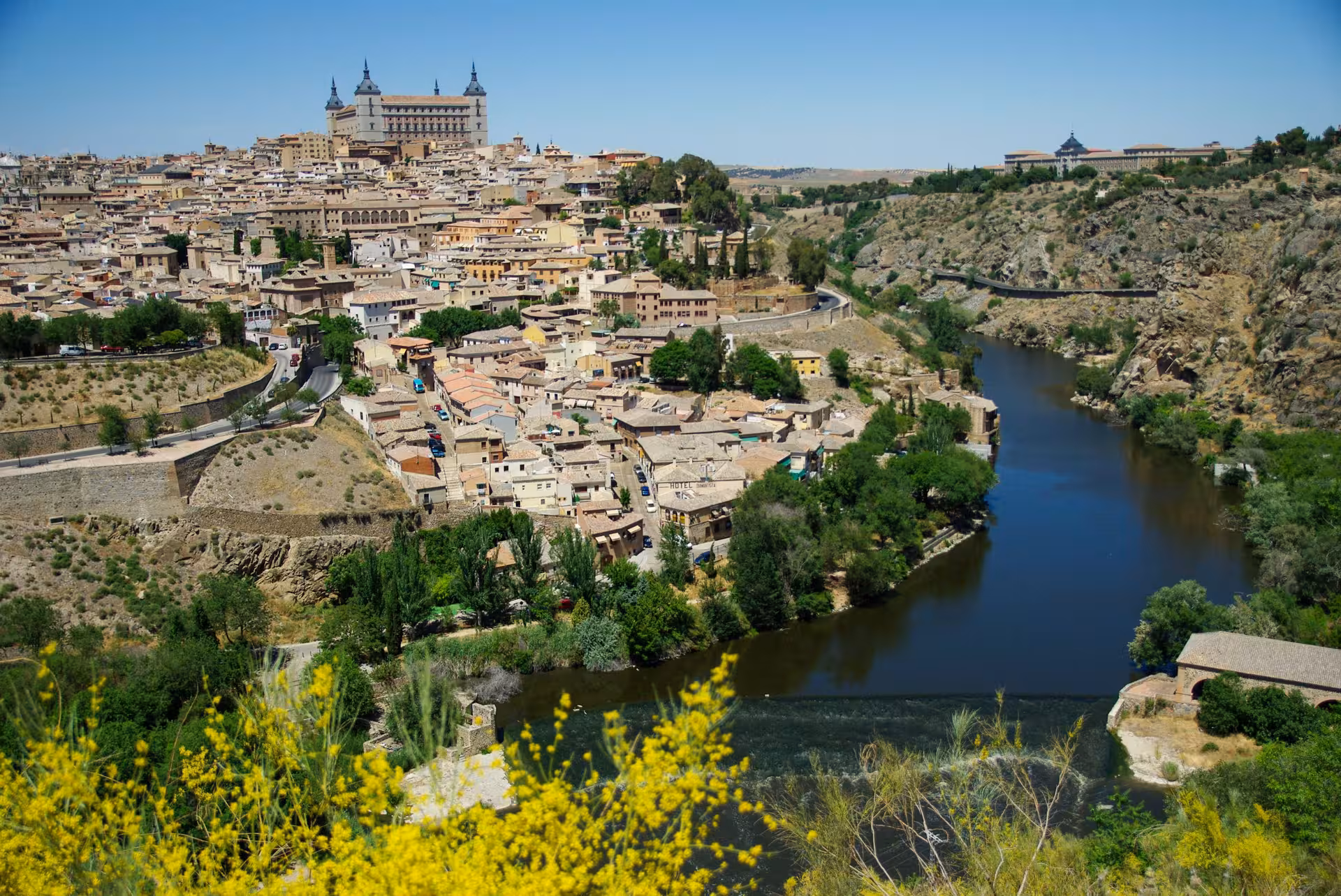 Panoramic view of Toledo cityscape, highlighting the Alcázar and winding Tagus River under a clear blue sky.