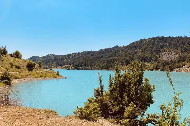 Scenic view of turquoise Tomor Mountain Lake surrounded by lush greenery under a clear blue sky in Dardhe, perfect for hiking and swimming tours.