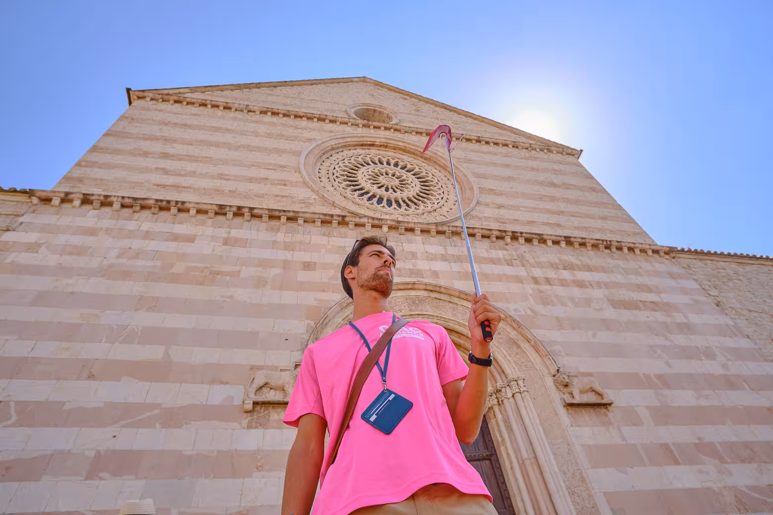 Tourist holding a selfie stick in front of Assisi's Basilica, capturing its intricate facade under the bright sun.