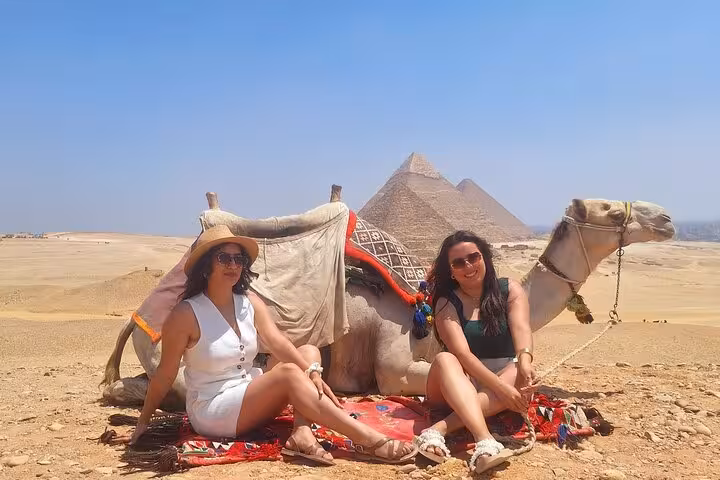 Two tourists sit with a camel in front of the iconic Giza Pyramids under a clear blue sky during a private Cairo tour.