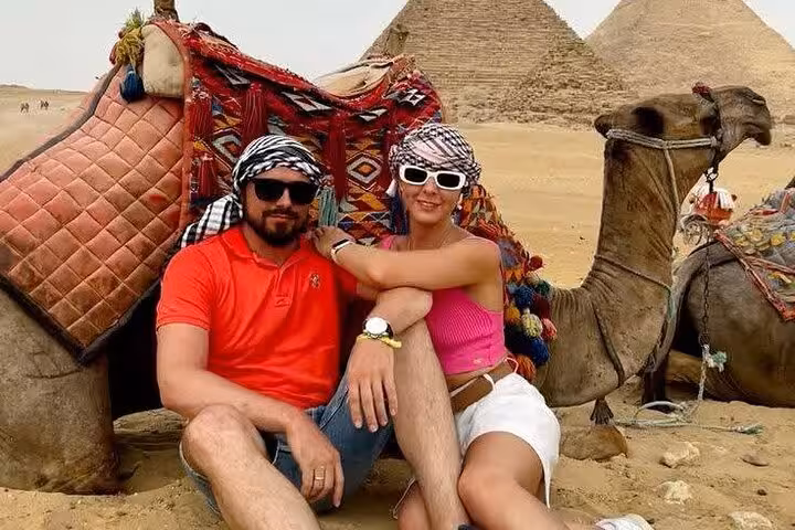 Tourists resting beside decorated camel with Giza Pyramids backdrop on private Giza tour with bazaar and museum