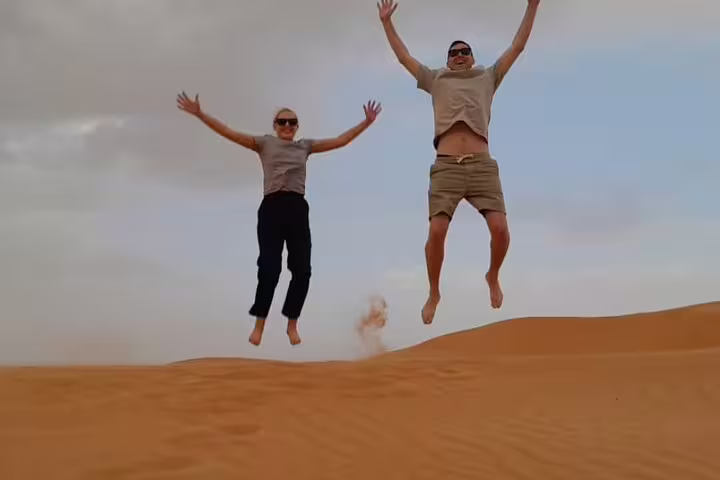 Two tourists joyfully jumping on golden sand dunes under a clear sky during a desert sunset watching experience.