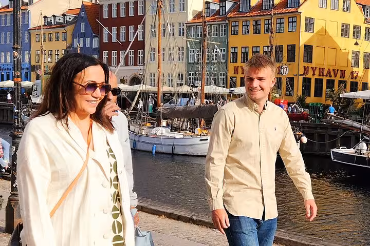 Smiling tourists stroll along the vibrant Nyhavn waterfront in Old Copenhagen, with colorful buildings and boats.