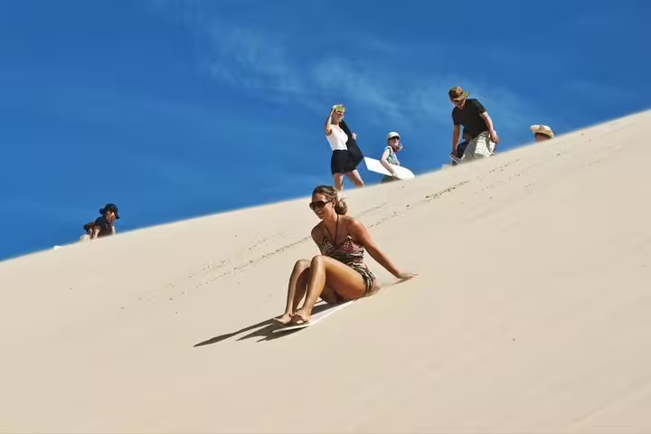 Tourists sandboarding down a steep desert dune, capturing the thrill of adventure against a vibrant blue sky.