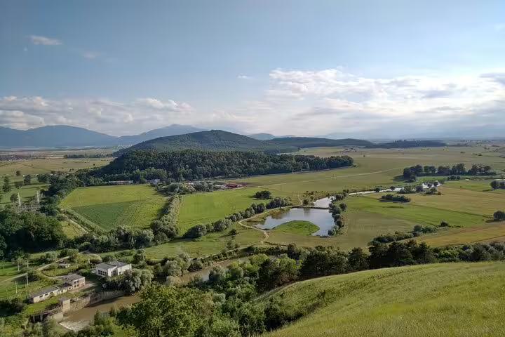 Aerial view of Transylvania valley with green fields, a small pond, forested hills, and mountains in the background