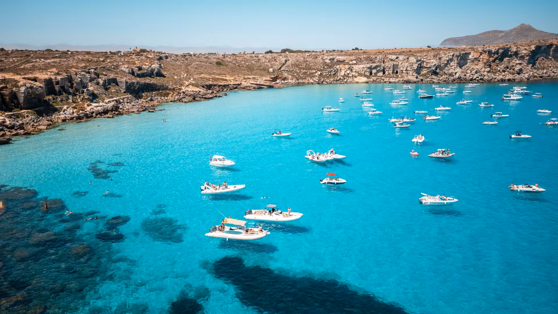 Aerial view of boats anchored in crystal-clear waters near rocky shores on a Trapani dinghy tour.
