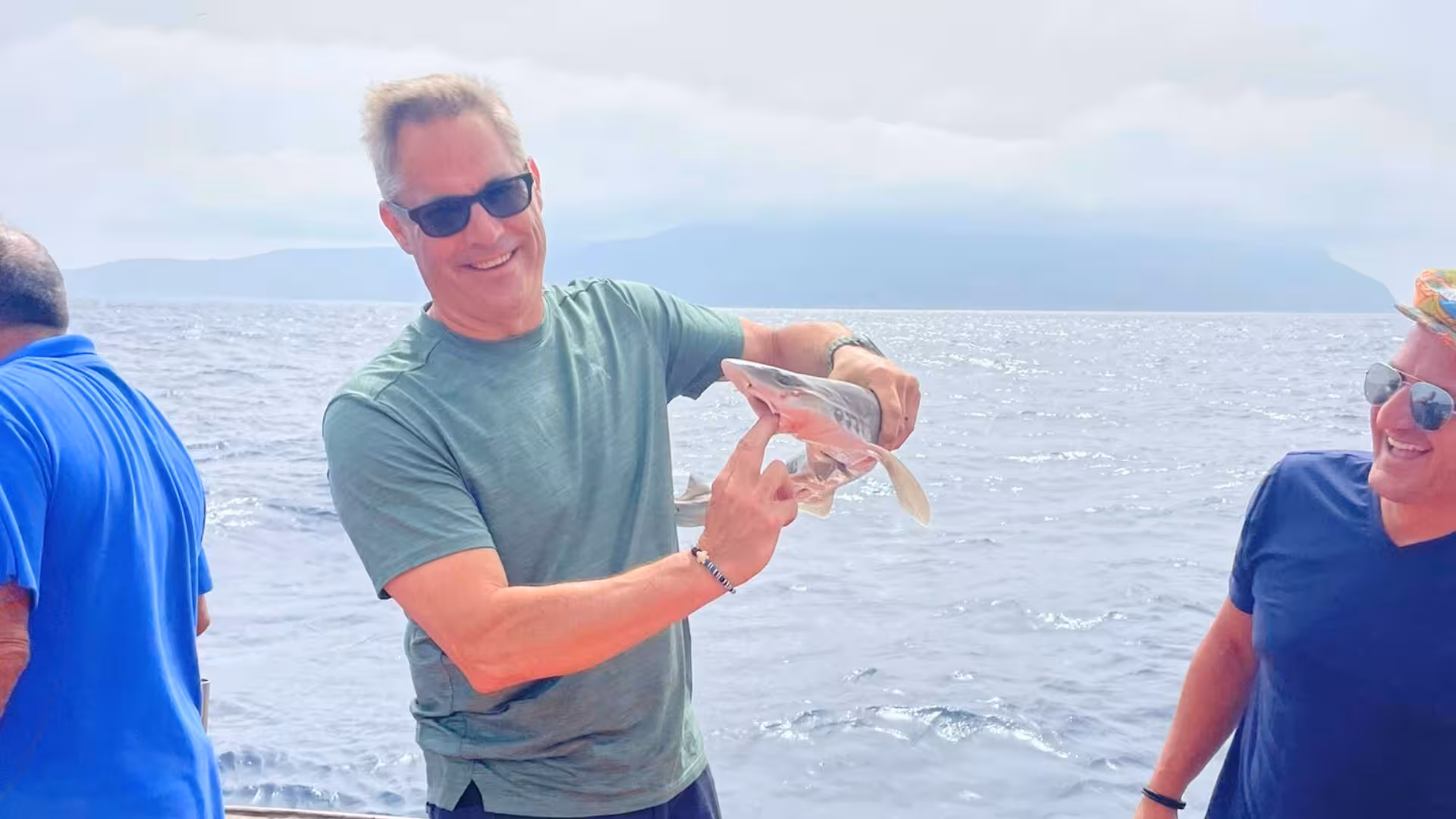 Tourist proudly holding a small fish on a Trapani fishing tour in the scenic Egadi Islands, with friends enjoying the moment.