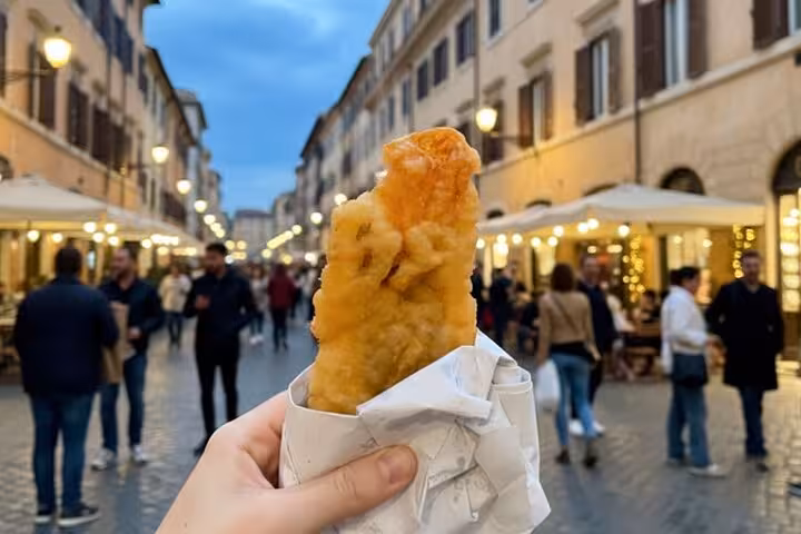 A hand holding a crispy Roman street food snack with a bustling Trastevere street in the background during a food tour.