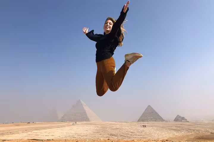 Traveler jumping at Giza Pyramids panoramic viewpoint during private tour, wide desert landscape and pyramids behind