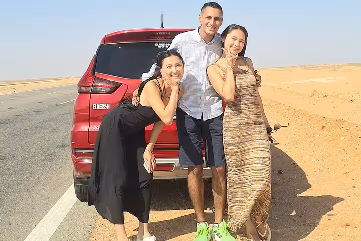 Smiling travelers pose beside a red SUV on a sunlit desert road, ready for an all-inclusive Black and White Desert adventure.