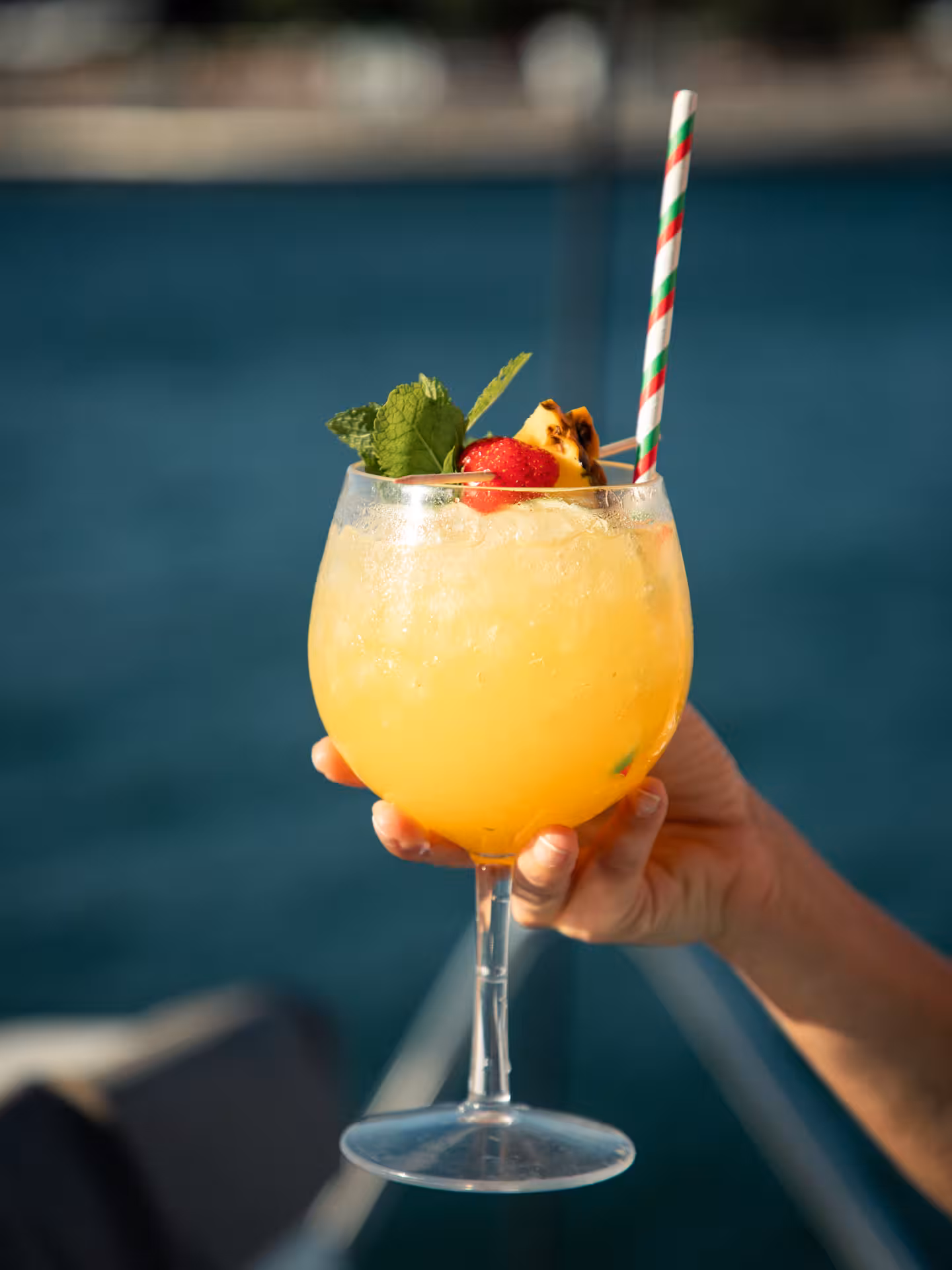 Hand holding tropical orange cocktail with mint, strawberry garnish and striped straw, cocktail masterclass drink