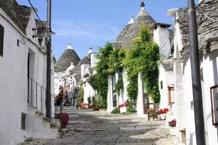 Sunlit street of whitewashed trulli houses with stone roofs and greenery in Alberobello, visited on a private tour from Naples