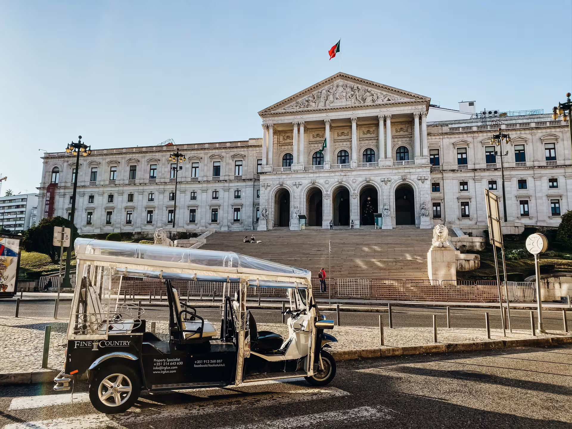 Tuktuk parked in front of Lisbon's historic parliament building under a clear blue sky.