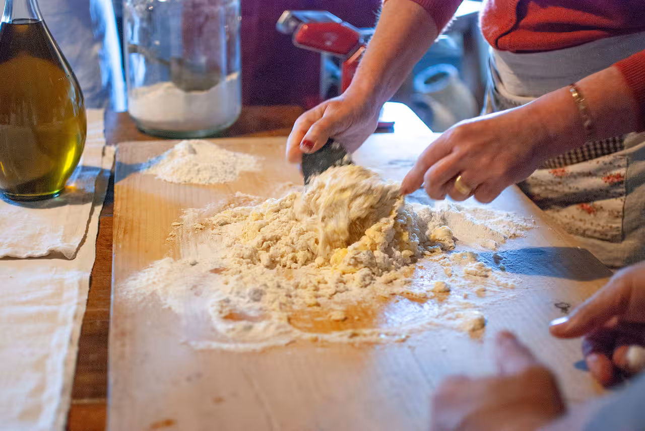 Hands mixing fresh dough on a wooden board during a Tuscany cooking course with olive oil and flour jars nearby.