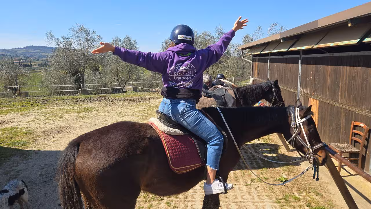 Person joyfully raising arms while riding a horse in scenic Tuscany countryside under a clear blue sky.