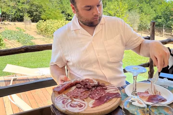 Man samples Italian cured meats on a wooden platter at an outdoor dining area in Tuscany's picturesque countryside.