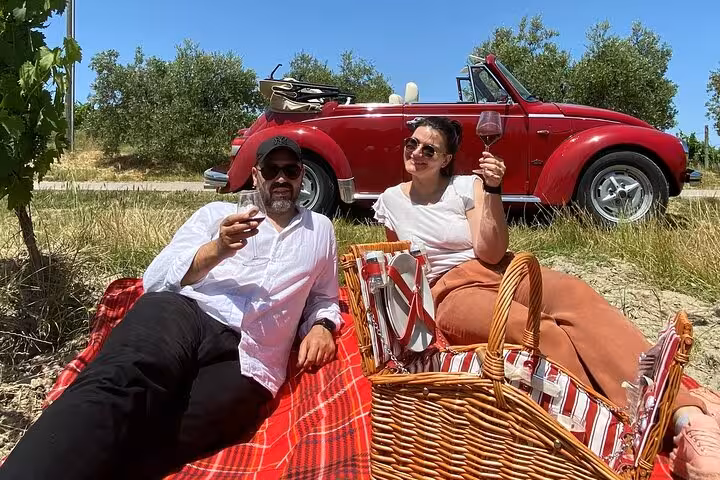 Couple enjoying a picnic with wine beside a red vintage Beetle in the scenic Tuscan countryside.