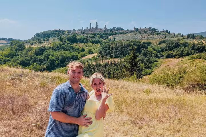 Excited couple on a Tuscany wine tour, with panoramic views of vineyards and historic architecture in the background.
