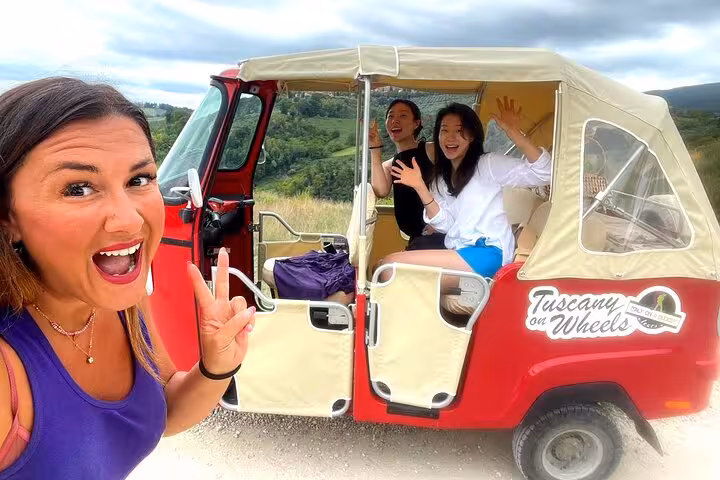 Guests smiling and posing with a red Tuk Tuk on a scenic Tuscany wine tour, surrounded by lush landscapes.