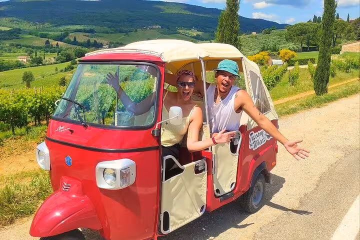 Couple enjoying a Tuscany wine tour in a red tuk tuk amidst scenic vineyards and rolling hills.