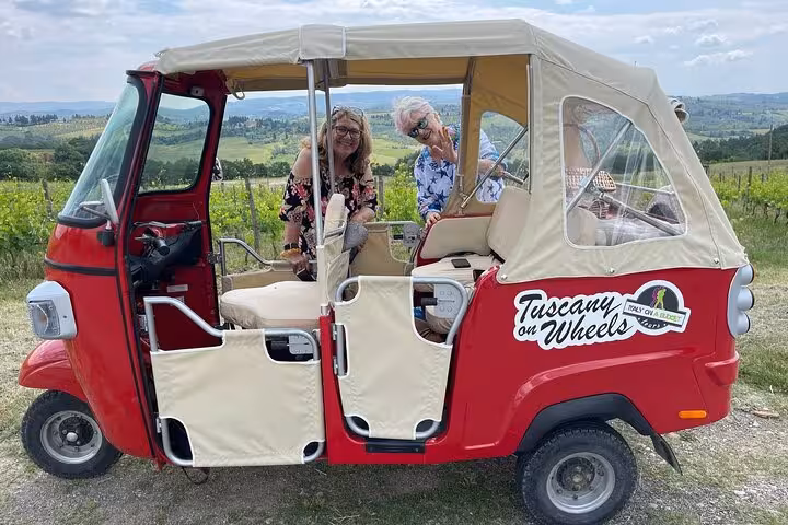 Two tourists ready for a Tuscany wine tour in a red tuk tuk with vineyard views.