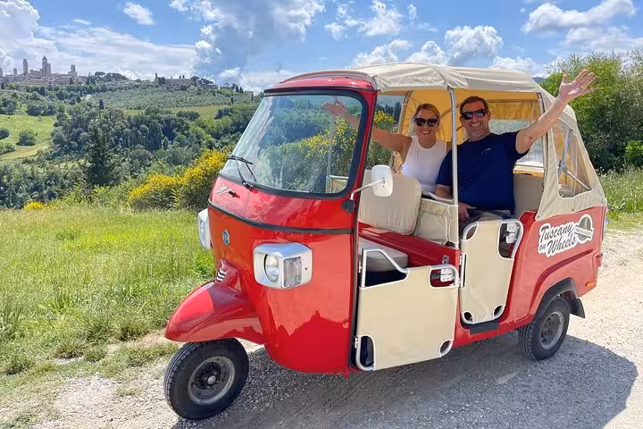 Couple enjoying Tuscany wine tour in a red tuk-tuk with scenic vineyard views under a bright blue sky.
