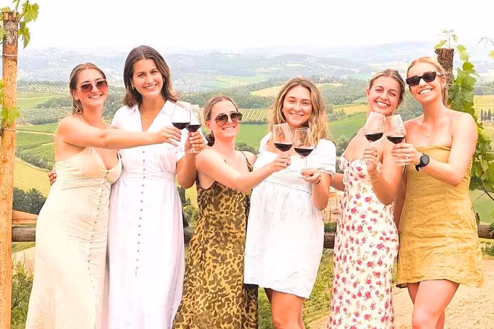 Six women raising wine glasses against the backdrop of Tuscany's rolling hills on a wine tour from Rome.