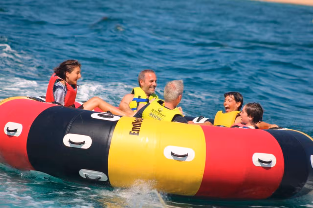 Group enjoying Twister inflatable tube ride, laughing together as they bounce over turquoise waves on a sunny beach day