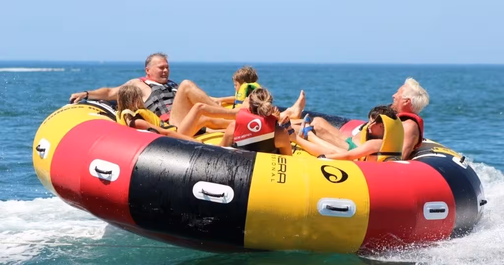 Group of friends laughing on a colorful Twister inflatable tube ride, speeding across clear blue sea on a sunny beach day