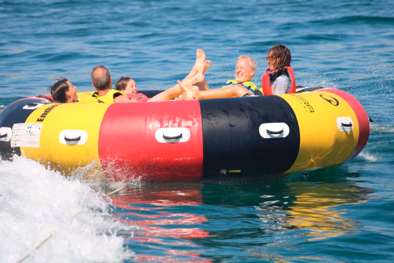 Friends on colorful Twister water tube, splashing and laughing while being towed fast across clear blue Mediterranean sea