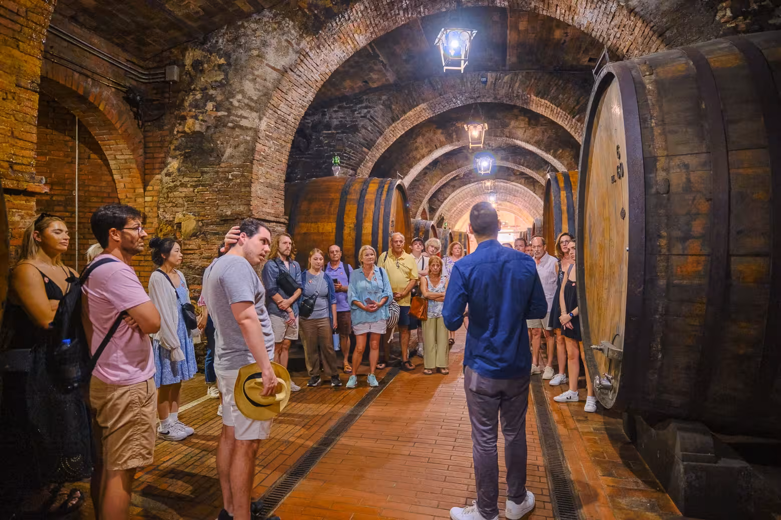 Tour group listening to a guide in a historic Val D'Orcia wine cellar with large wooden barrels, perfect for wine tasting tours.