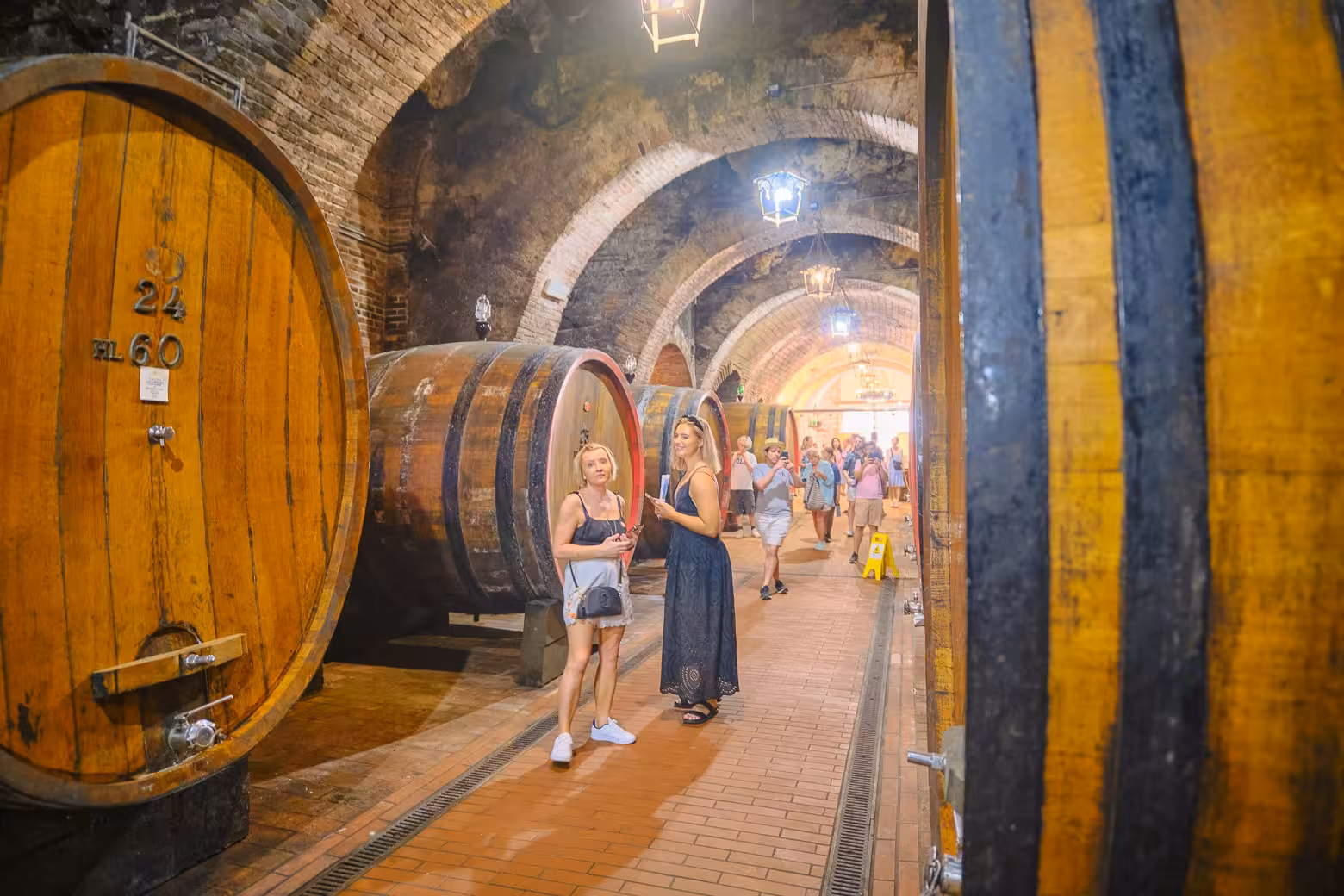 Tourists exploring a historic wine cellar in Val D'Orcia with large wooden barrels lining the brick-vaulted hall.