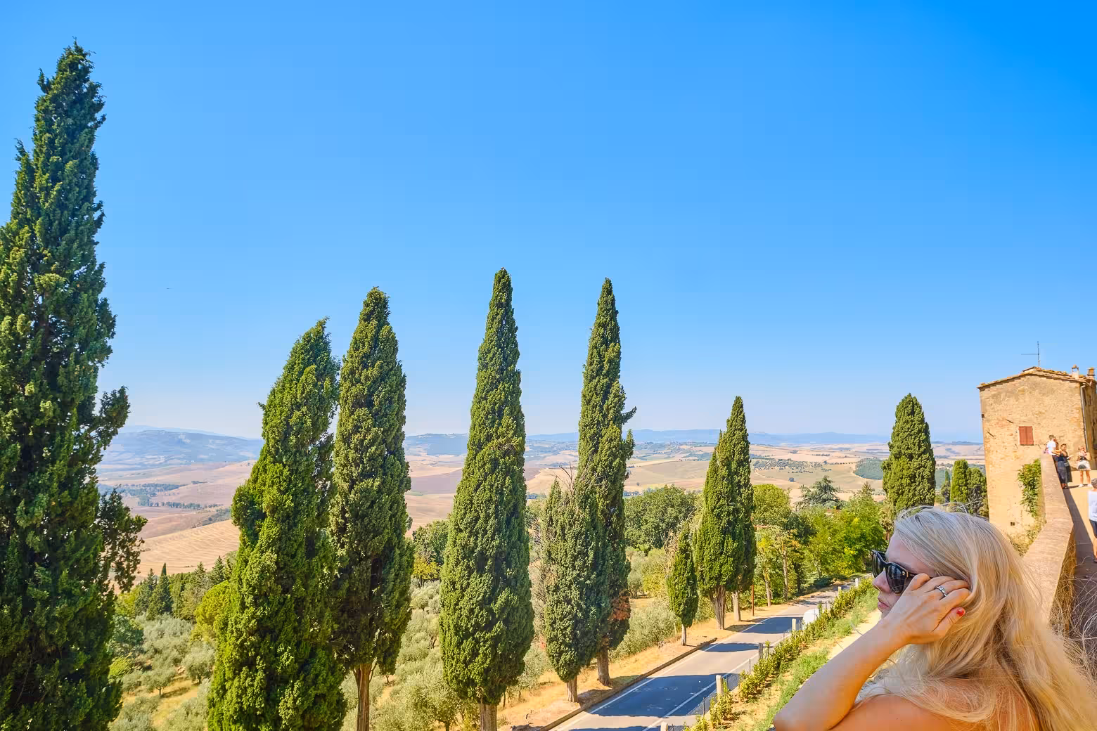 A visitor enjoys the panoramic view of Val D'Orcia's landscape, framed by iconic cypress trees and sunny skies.
