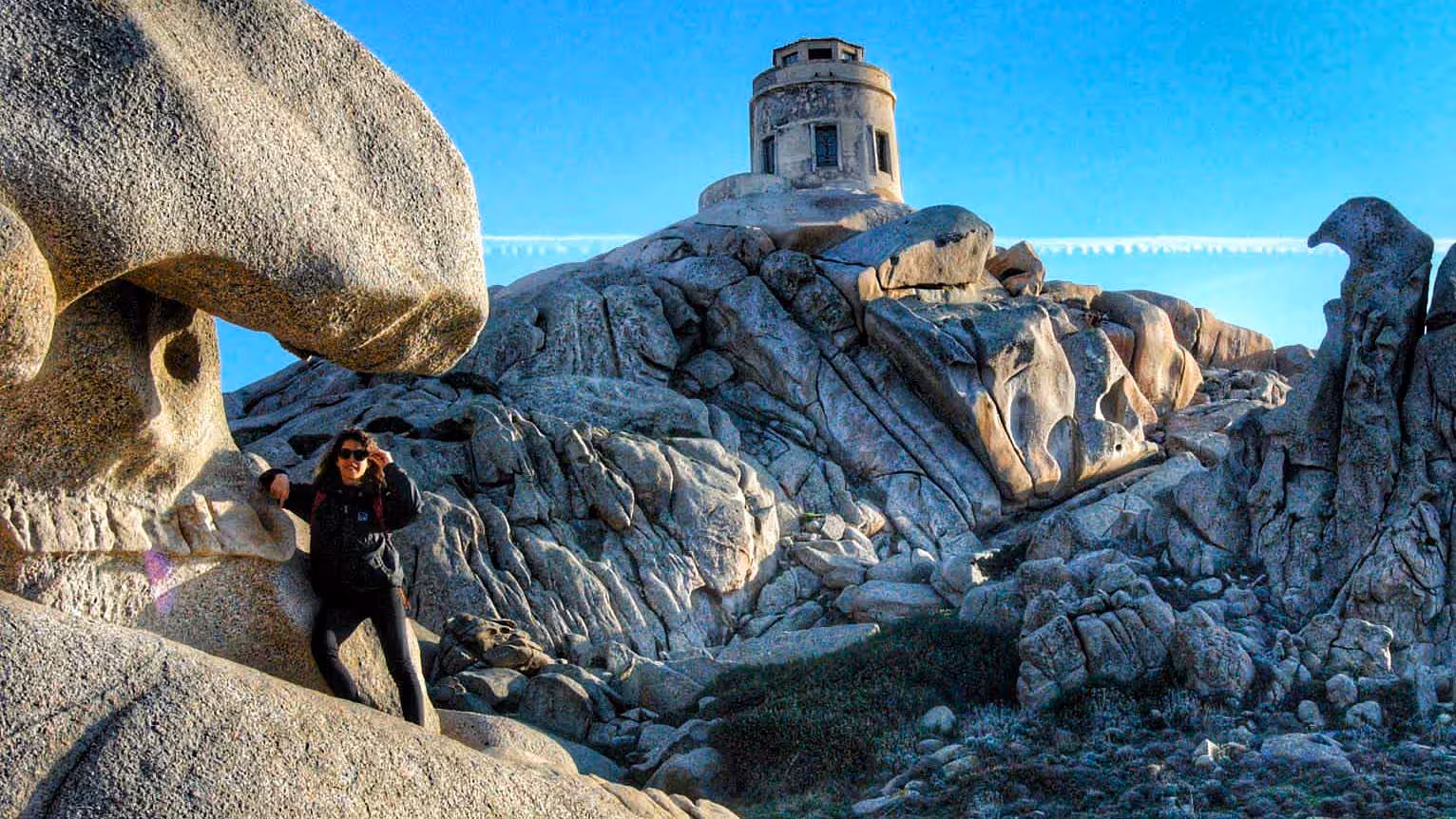 Hiker exploring unique rock formations in Valle della Luna with a historic tower in Santa Teresa Gallura, Sardinia.