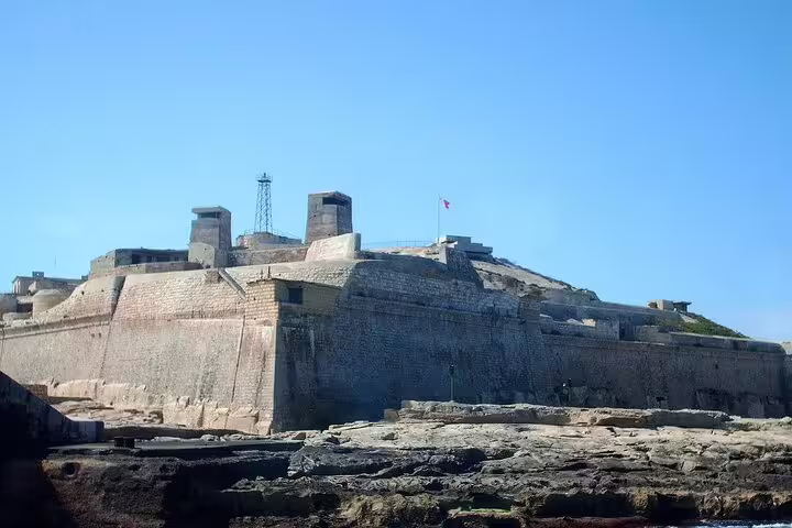 Majestic stone fortification with a flag atop, overlooking the rocky coastline in Valletta's harbor area.