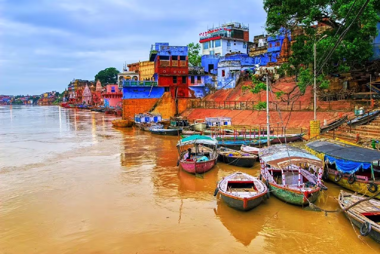 Colorful ghats of Varanasi along the Ganges River with moored boats and vibrant buildings under a cloudy sky.