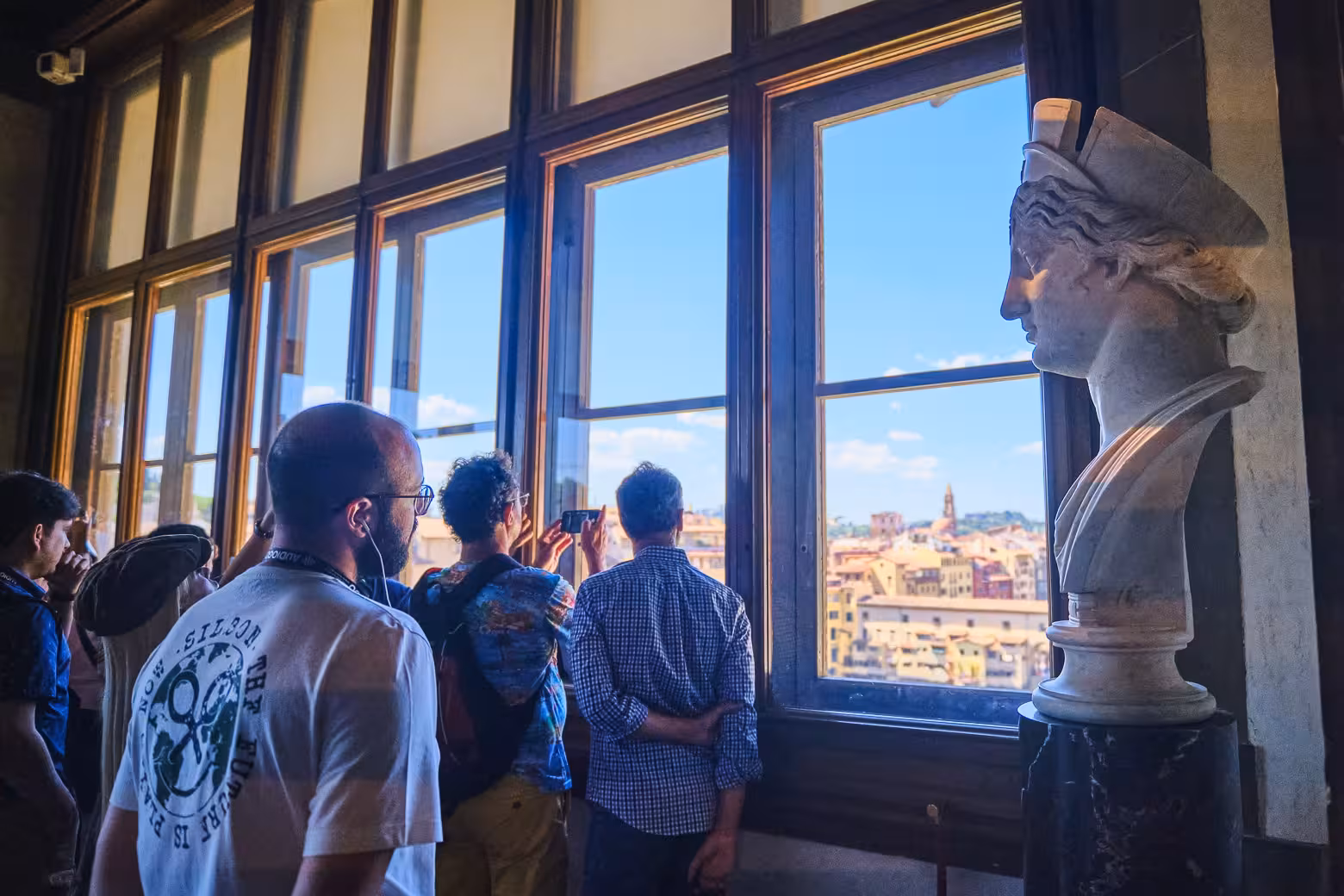 Visitors in Vasari Corridor admiring Florence views through large windows, next to classical sculptures.
