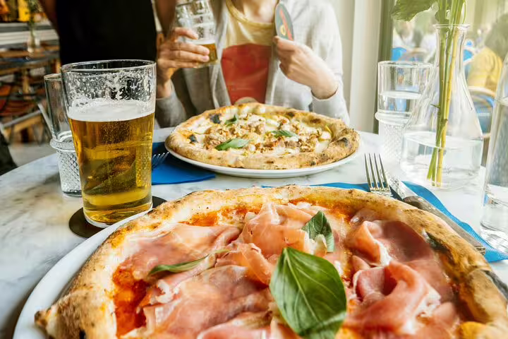 Two gourmet pizzas with fresh toppings and glasses of beer on a table, capturing the essence of the Vatican Market Experience tasting tour.