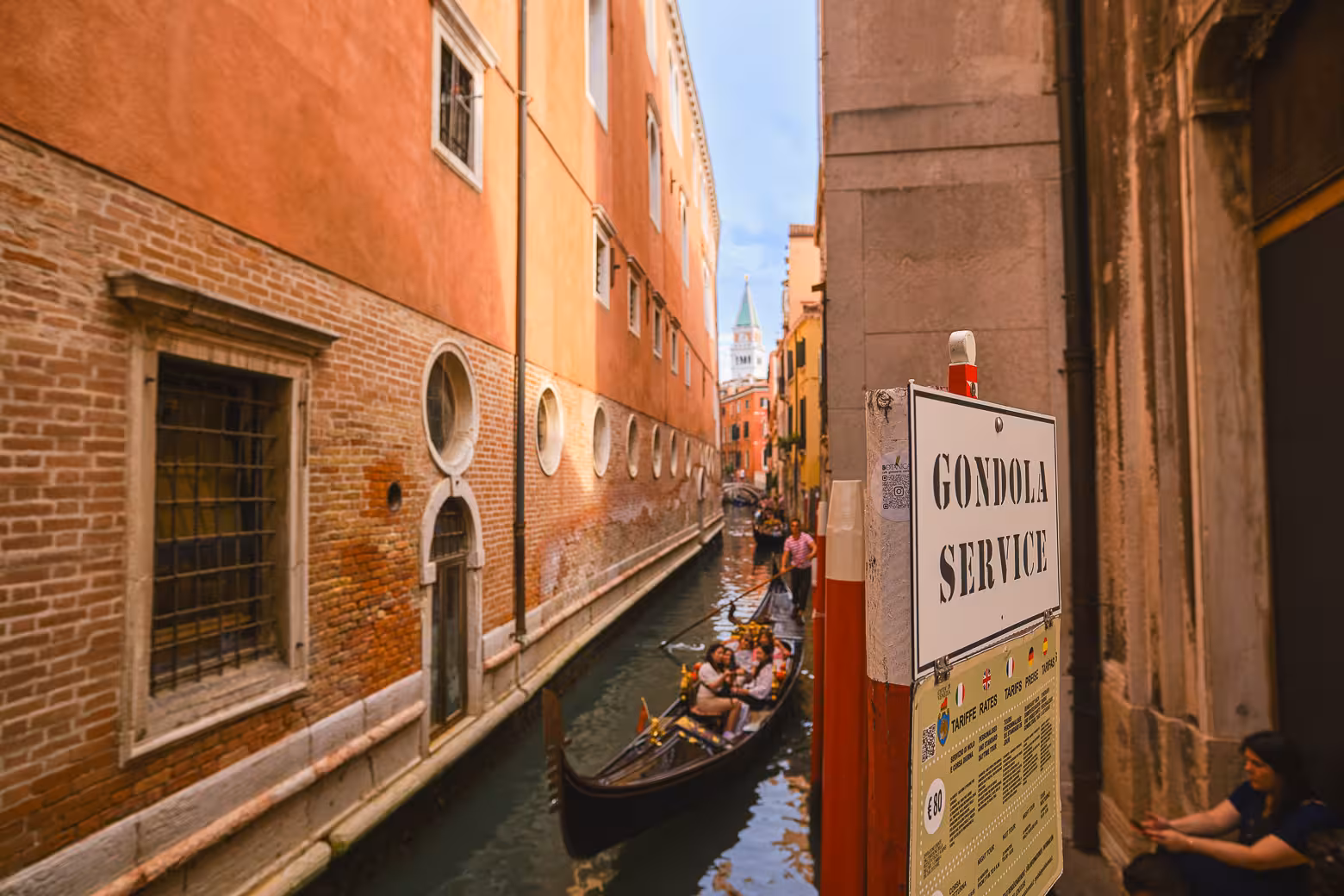 Venice gondola service sign by a narrow canal with tourists enjoying a classic gondola ride through historic streets.