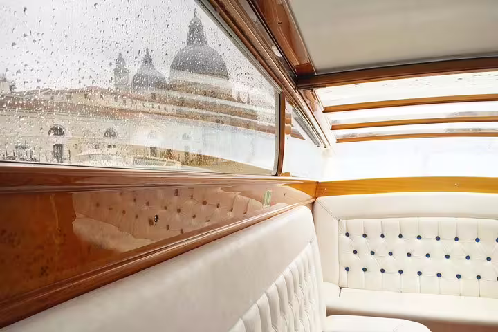 Rain-soaked window view from a private Venetian boat with plush seating and a glimpse of iconic architecture.