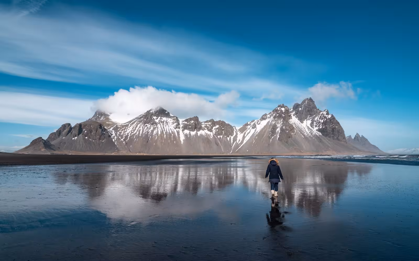 Person explores the reflective sands of Vestrahorn beach with dramatic snow-capped peaks on Iceland's South Coast.