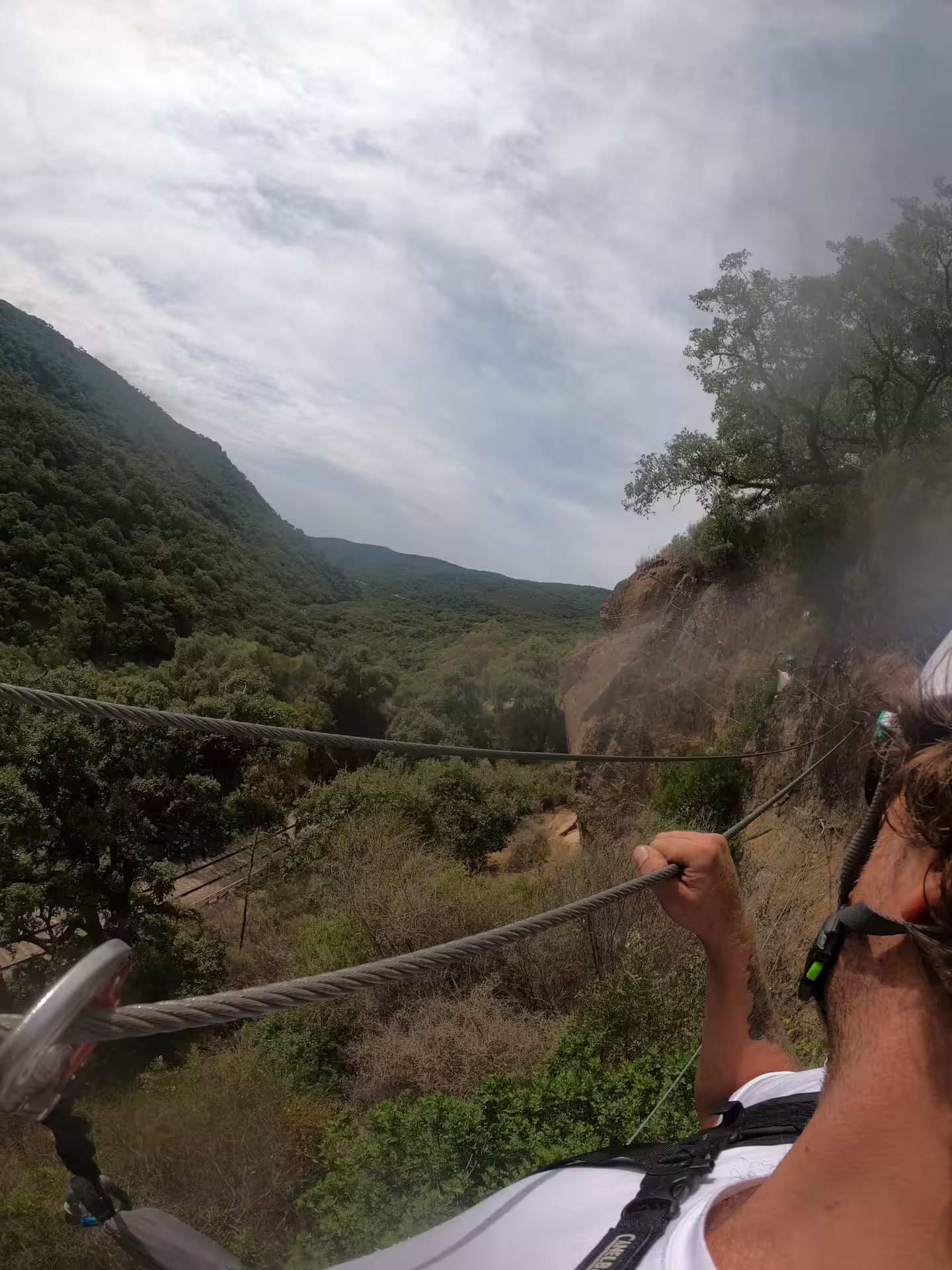 Thrill-seeker crosses cable bridge at Via Ferrata El Caimán, overlooking expansive views of Alcornocales' rolling hills.
