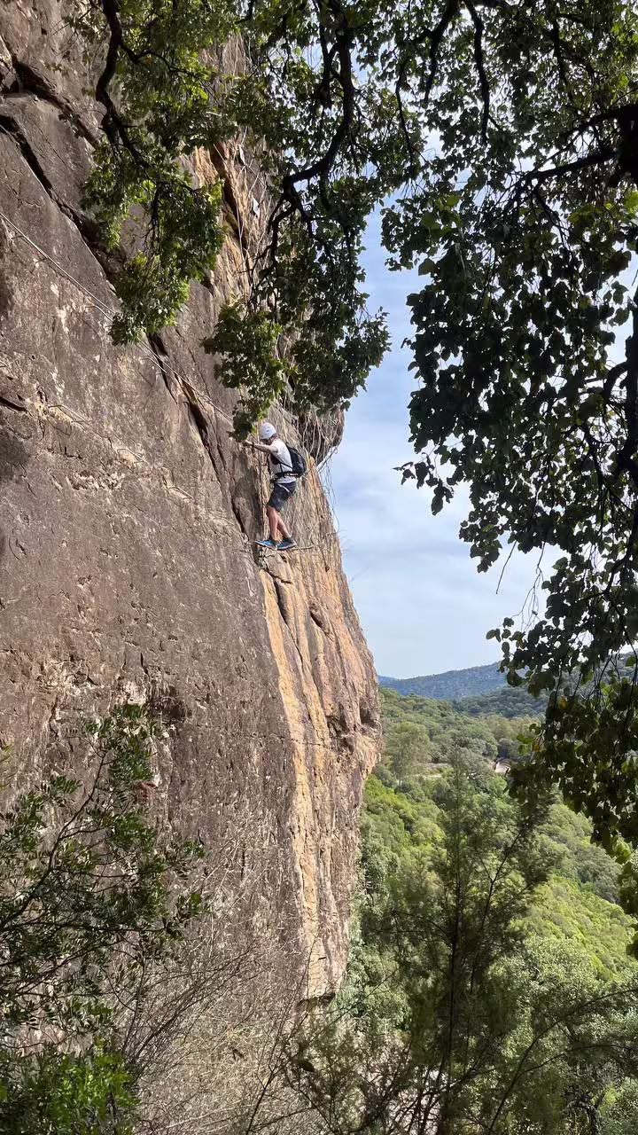 Climber navigating a steep rock face on Via Ferrata El Caimán in Alcornocales Natural Park, surrounded by lush greenery.