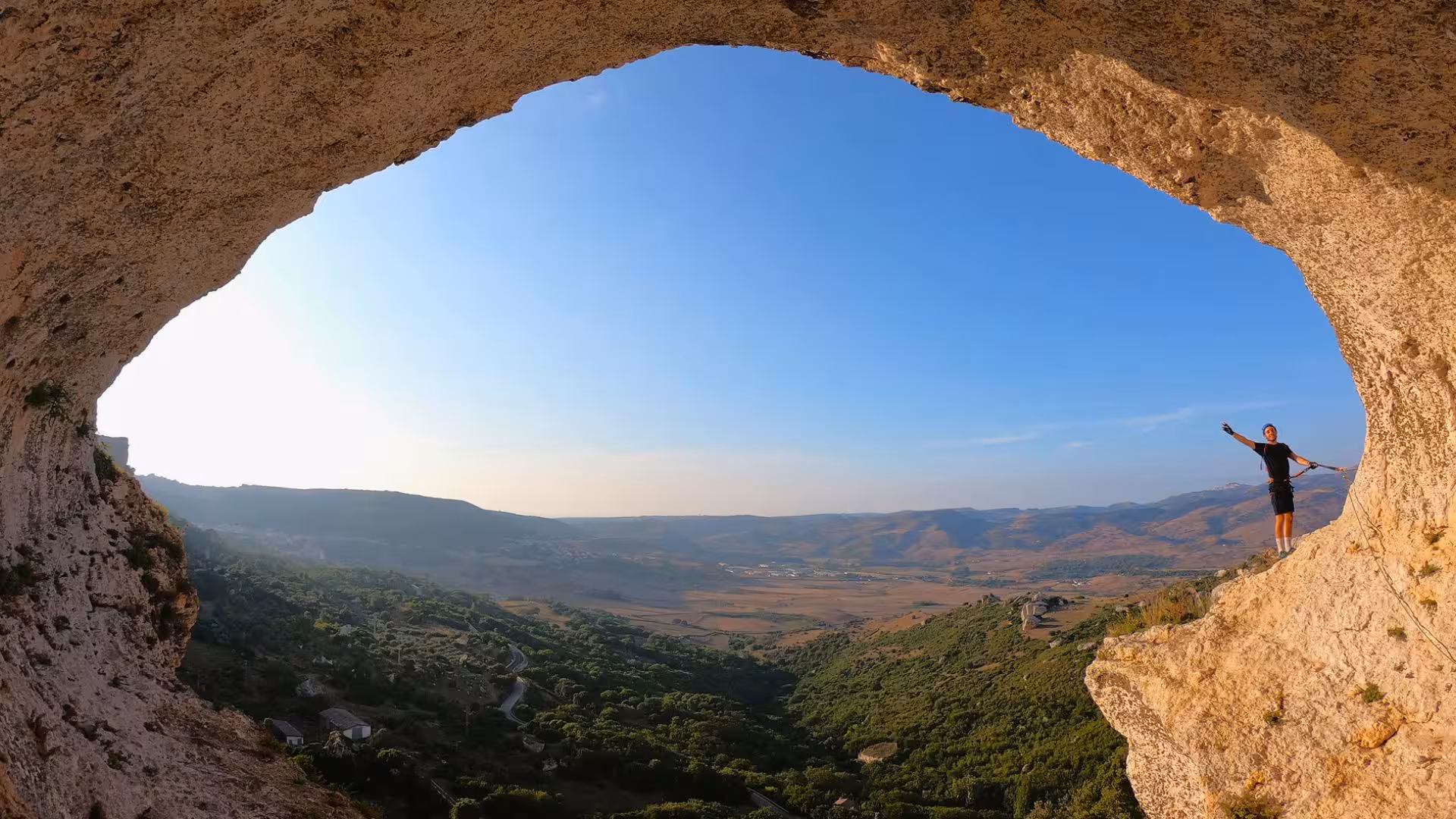 Explorer stands triumphantly under a rock arch on Via Ferrata di Giorré with panoramic views of Cargeghe's hills.