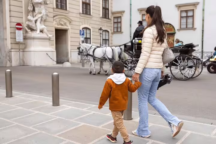 Family exploring Vienna on a private tour, passing horse-drawn carriages near historic city landmarks and elegant architecture.