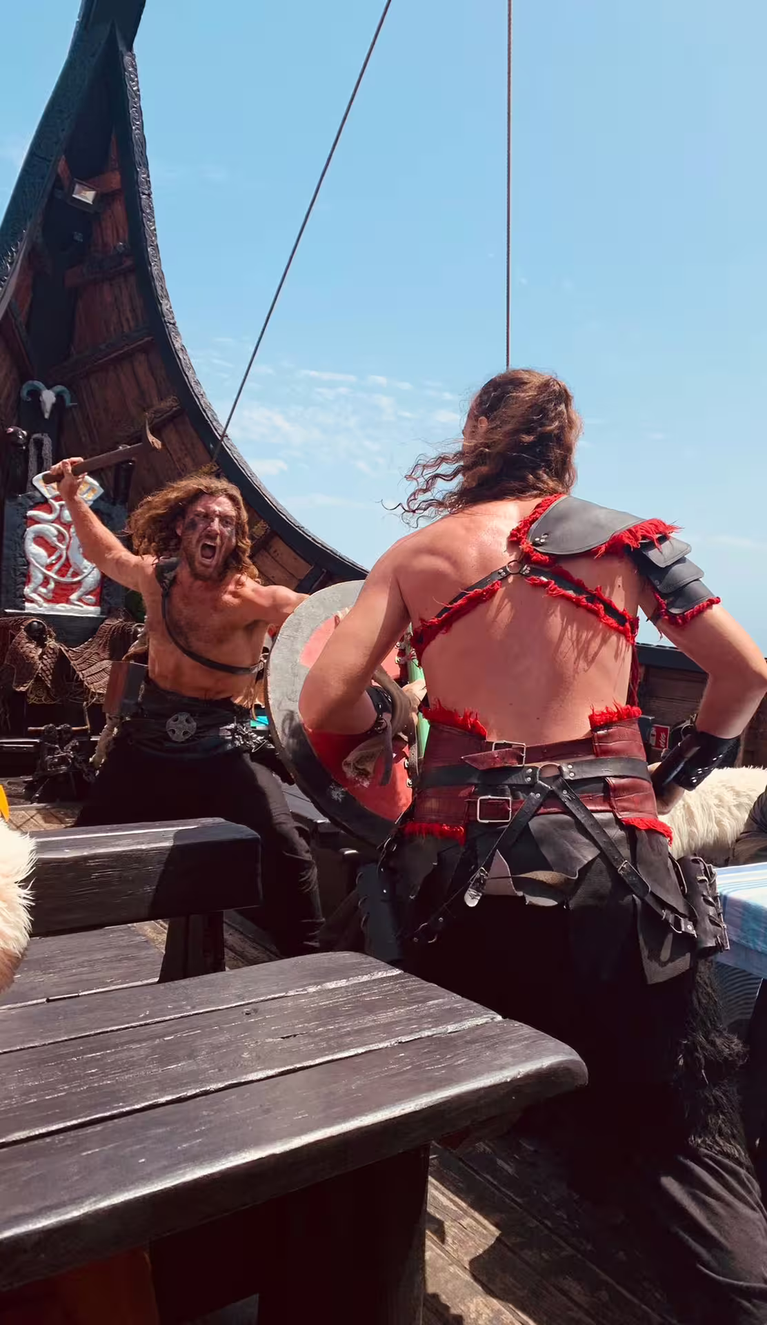 Viking reenactors in costume engage in a lively battle on a wooden ship under a clear blue sky.