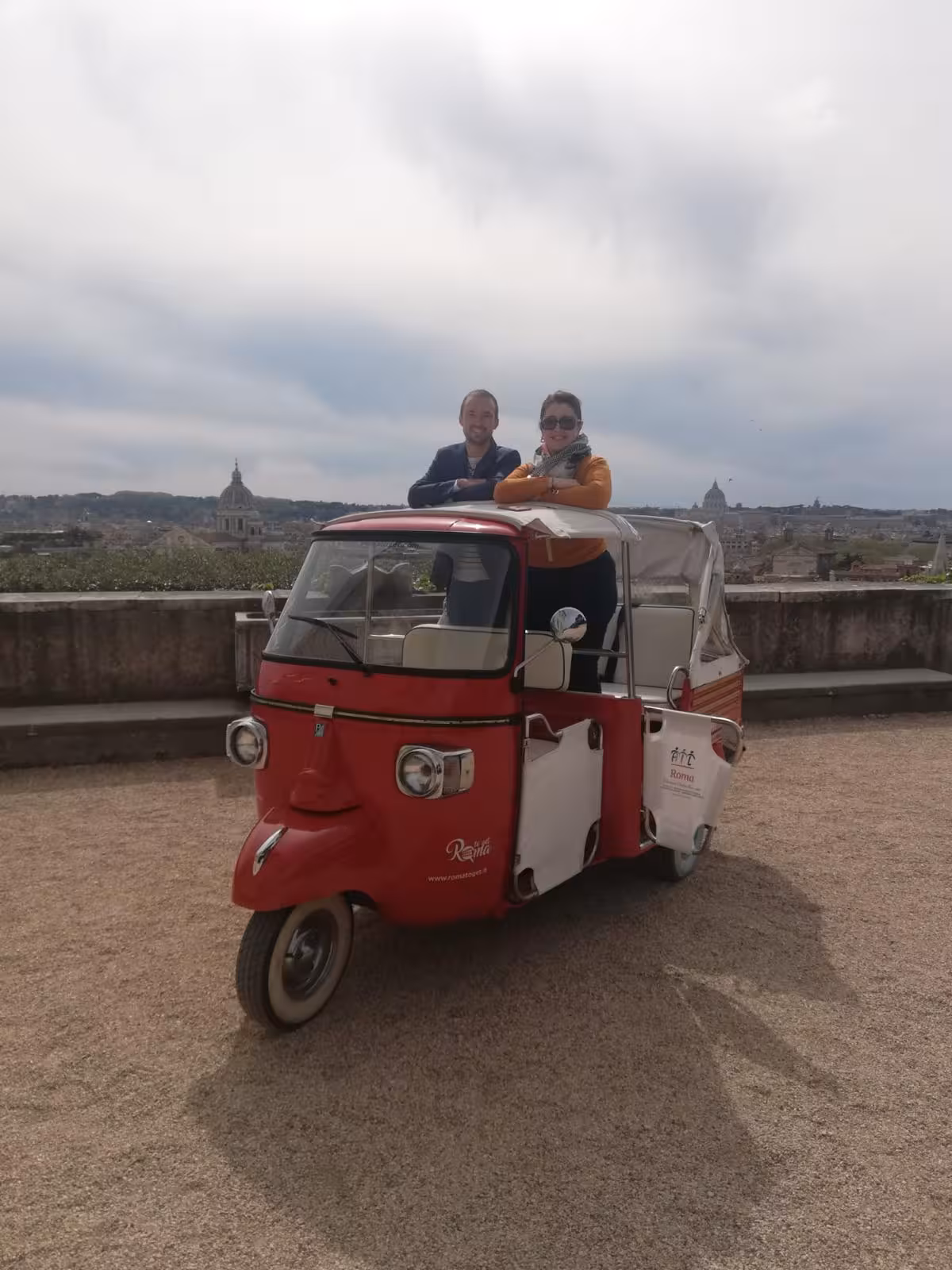 Two people standing in a red tuk-tuk with a scenic view of Rome in the background during Villa Medici tour.
