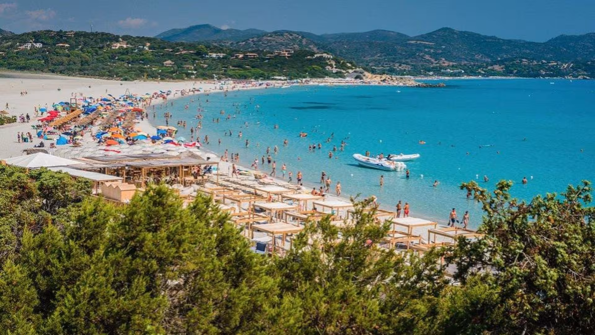 Crowded Villasimius beach with colorful umbrellas and a dinghy on turquoise waters, perfect for summer tours.