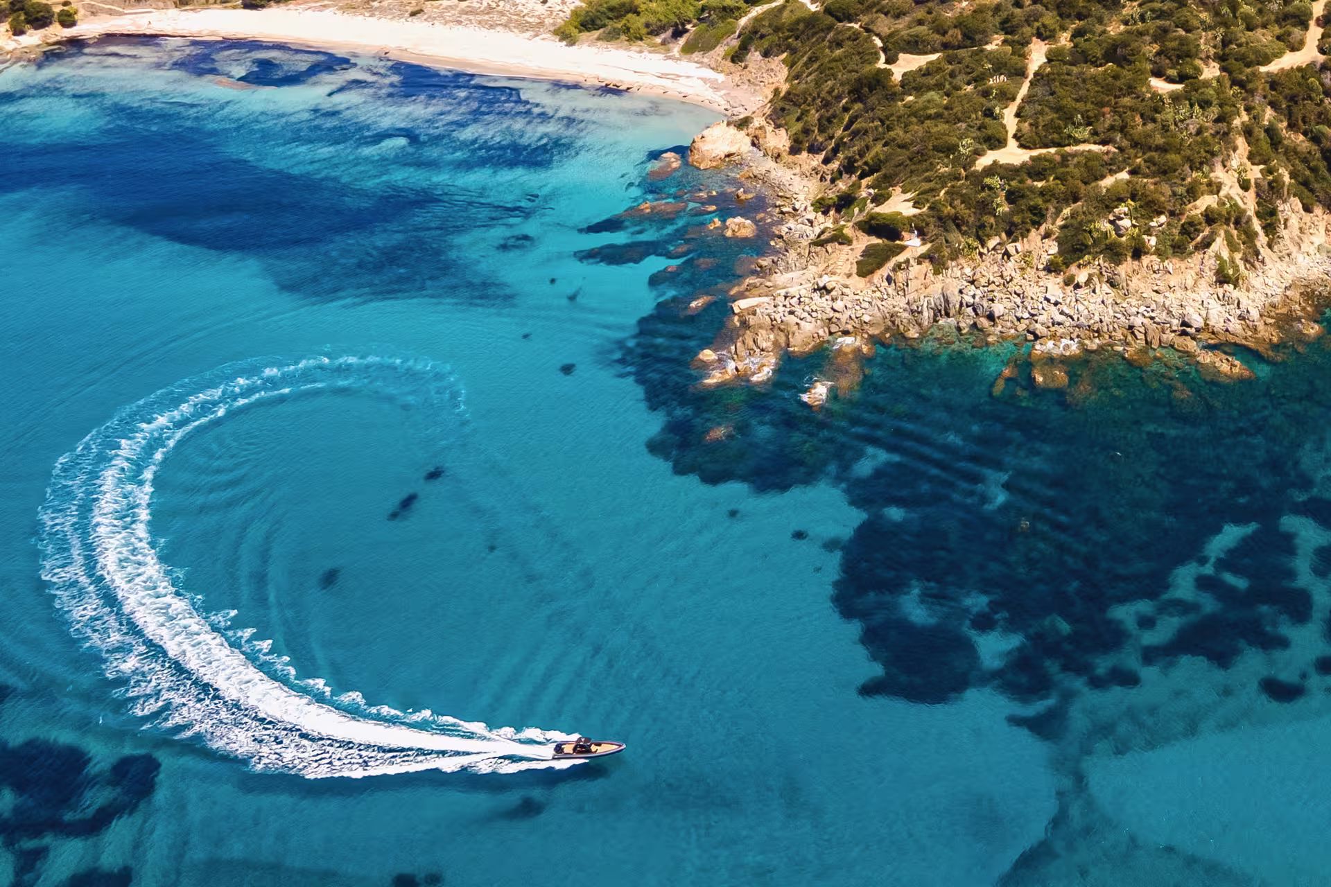 Aerial view of a RIB boat making a sweeping turn in the crystal-clear turquoise waters of Villasimius coastline.