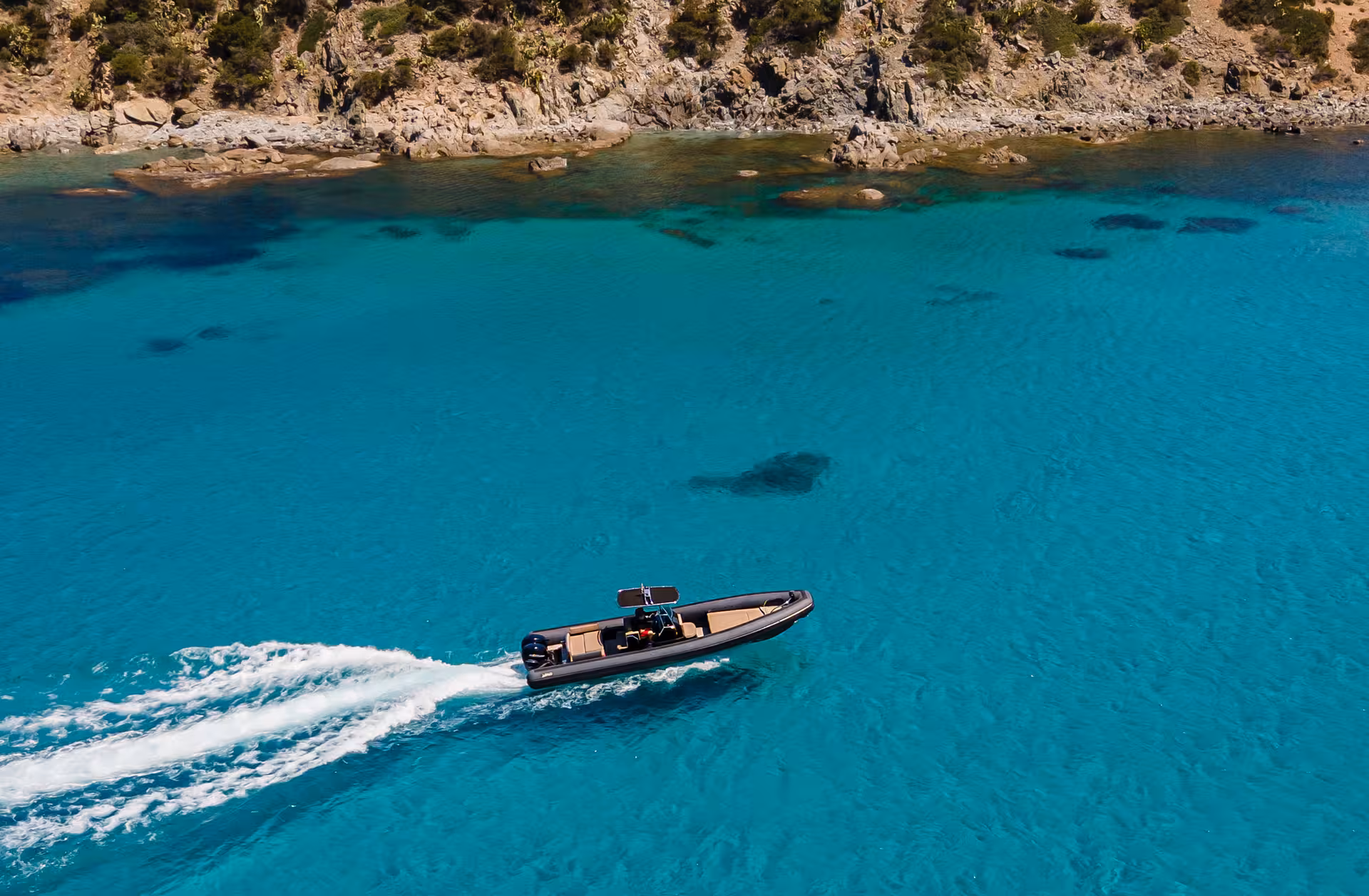 Aerial view of a RIB speeding through clear turquoise waters near rocky coastline in Villasimius tour.